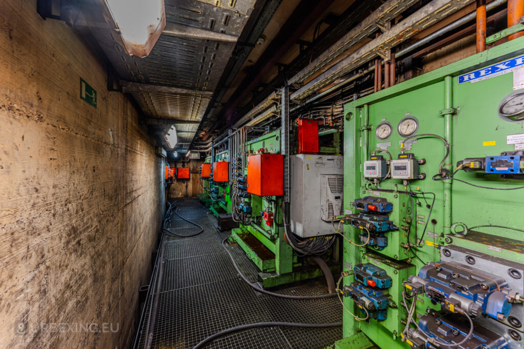 Long corridor in the abandoned Vallourec steel mill in Düsseldorf-Rath, featuring green machinery panels, bright orange control boxes, and exposed pipes along the walls and ceiling.