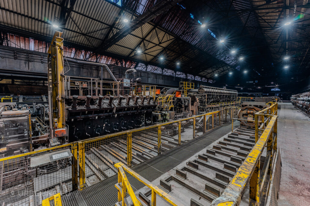 Detailed view of heavy industrial machinery in the abandoned Vallourec steel mill in Düsseldorf-Rath, with yellow safety railings and a high vaulted roof illuminated by factory lights.