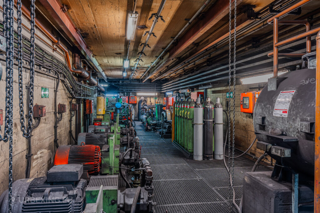 Industrial basement room in the abandoned Vallourec steel mill in Düsseldorf-Rath, featuring green machinery, gas cylinders, and exposed pipes and chains under fluorescent lighting.