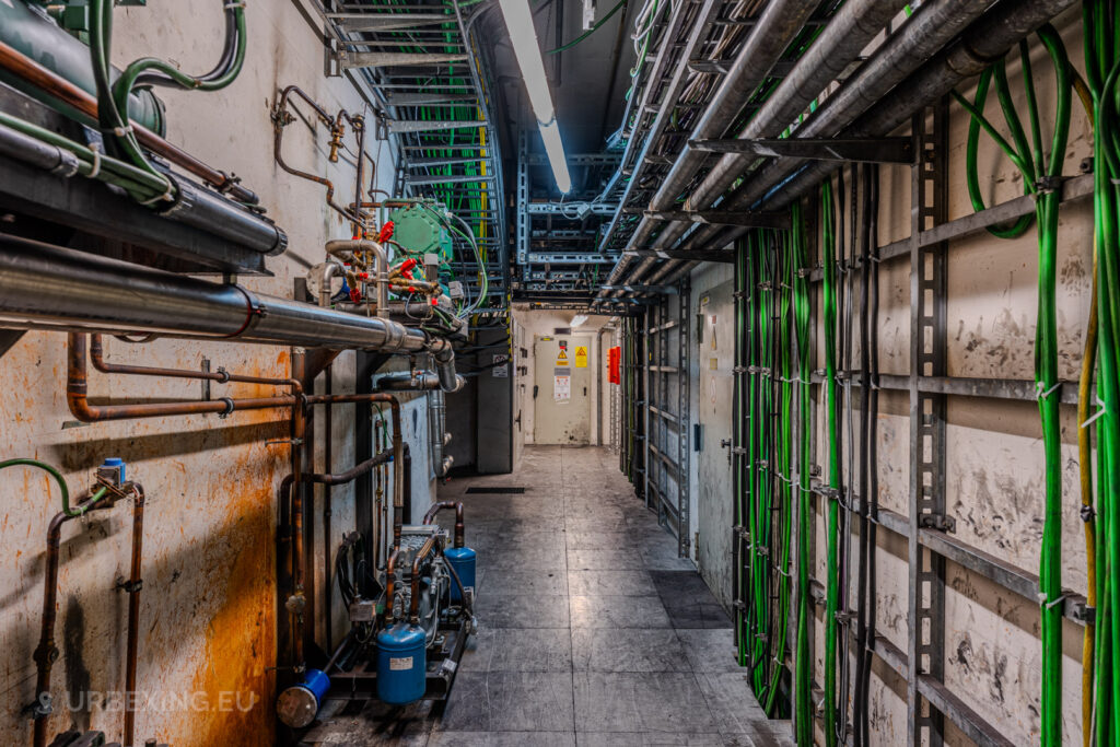 Corridor in the Vallourec steel mill in Düsseldorf-Rath, with exposed green and metallic pipes, industrial cables, and warning signs on a distant door.