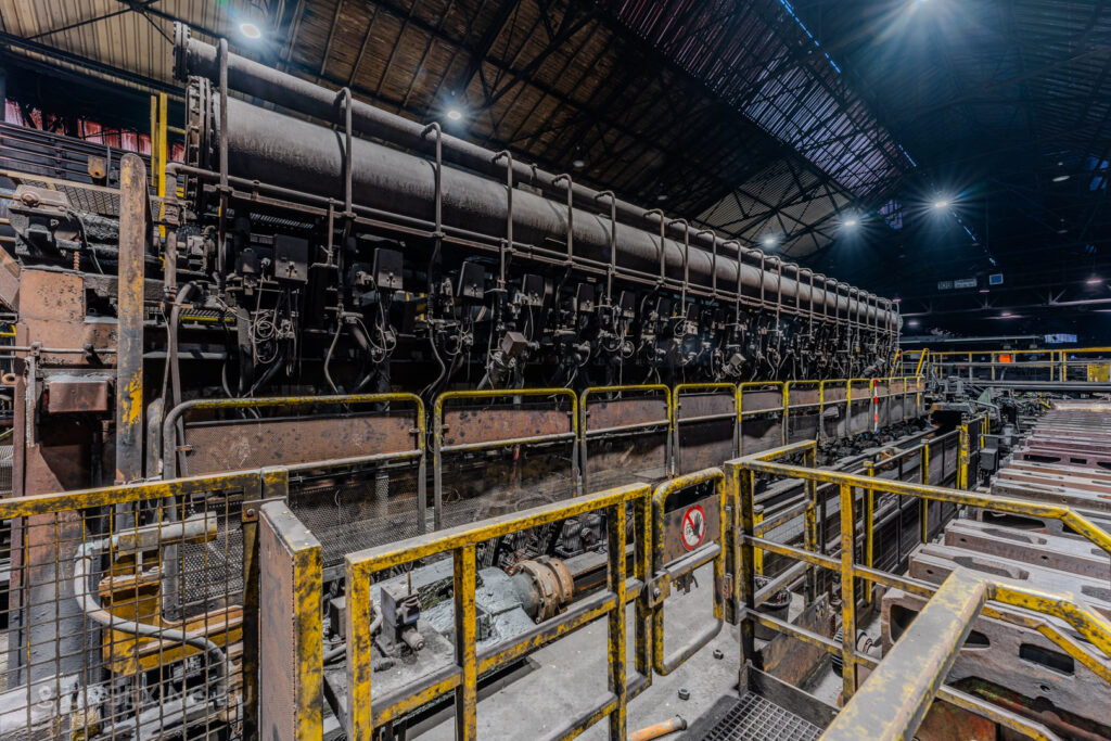 Close-up of a massive industrial roller machine in the abandoned Vallourec steel mill in Düsseldorf-Rath, framed by yellow railings and surrounded by signs of rust and decay.