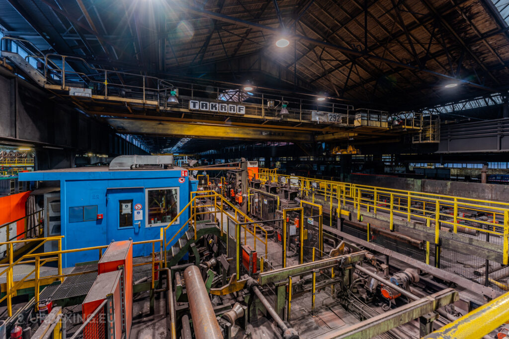 Industrial setup in the abandoned Vallourec steel mill in Düsseldorf-Rath, featuring a bright blue operator cabin, yellow safety railings, and an overhead TAKRAF crane in a vast manufacturing hall.