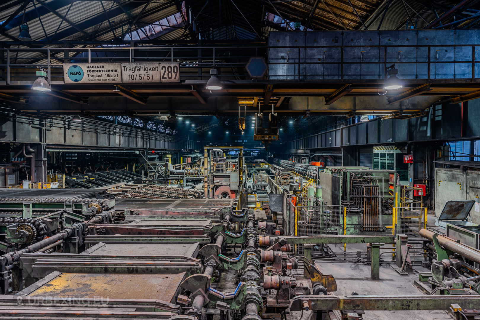 Close-up of the abandoned machinery and conveyor systems inside the Vallourec steel mill in Düsseldorf-Rath, with industrial decay and a weathered overhead crane under the vaulted roof.