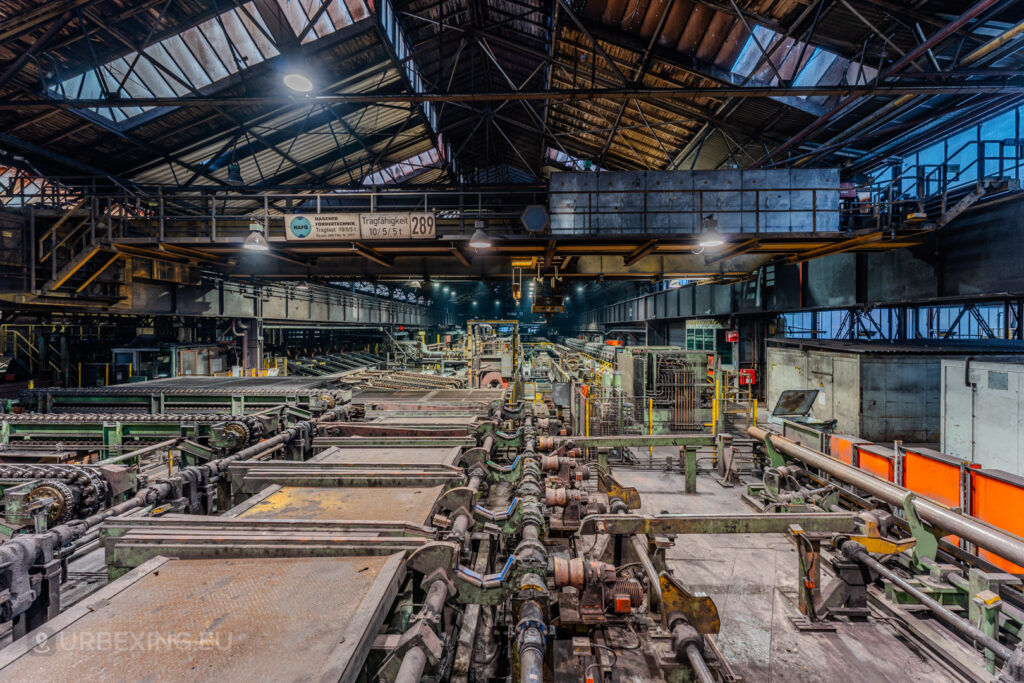 Extensive machinery and conveyor systems inside the abandoned Vallourec steel mill in Düsseldorf-Rath, with overhead walkways and industrial decay under a high vaulted roof.