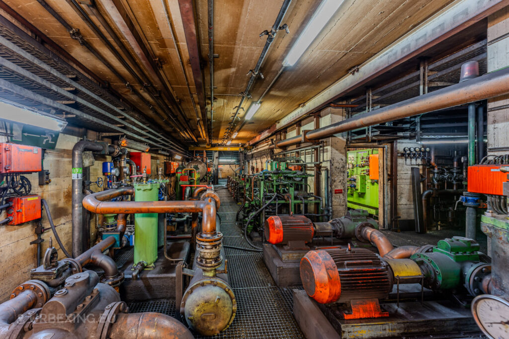 Underground mechanical room in the abandoned Vallourec steel mill in Düsseldorf-Rath, featuring rusted pipes, vibrant machinery, and exposed wiring illuminated by fluorescent lights.