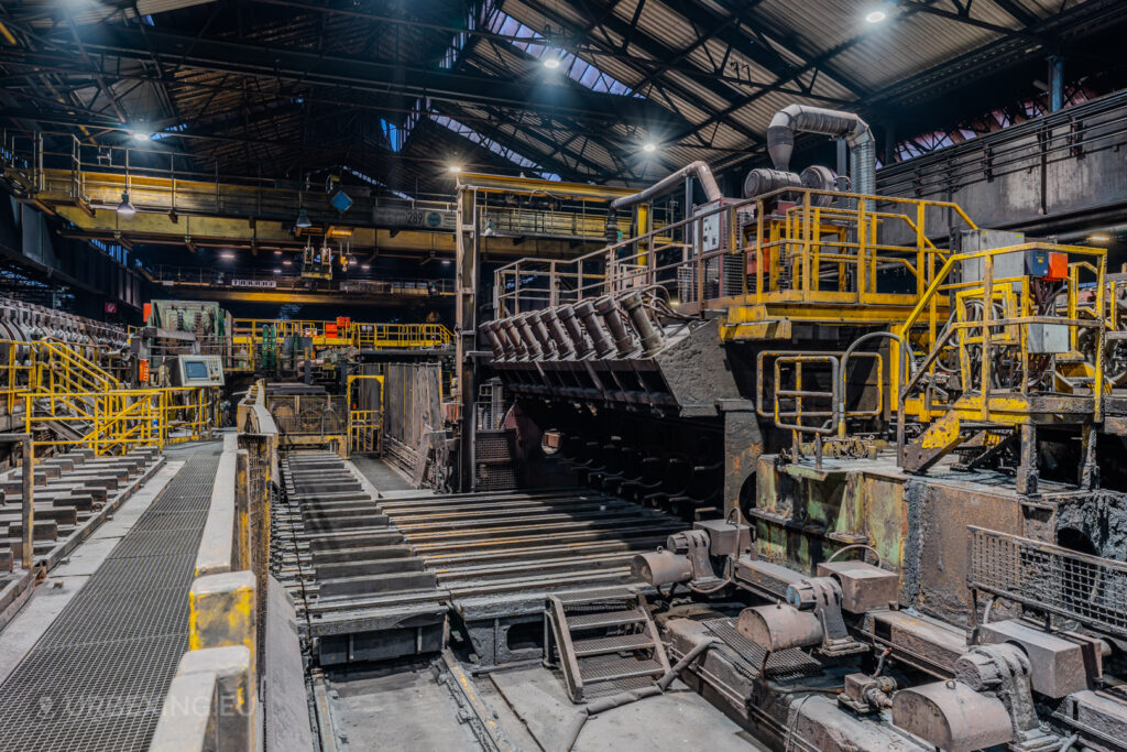 Industrial processing machinery in the Vallourec steel mill in Düsseldorf-Rath, surrounded by yellow railings and conveyor systems under a high arched roof.