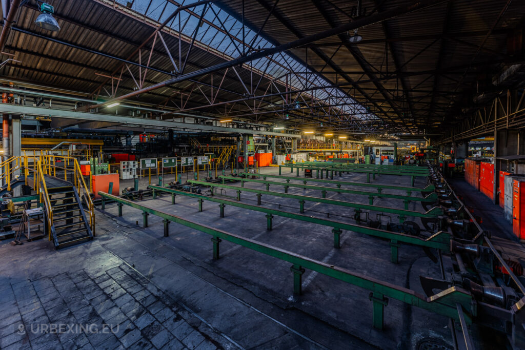 Wide-angle view of the production floor in the abandoned Vallourec steel mill in Düsseldorf-Rath, featuring green conveyor structures, red storage cabinets, and industrial walkways under a vaulted roof.