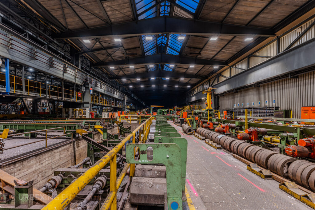 Expansive production line in the abandoned Vallourec steel mill in Düsseldorf-Rath, featuring green machinery, yellow railings, and a high arched ceiling illuminated by skylights.