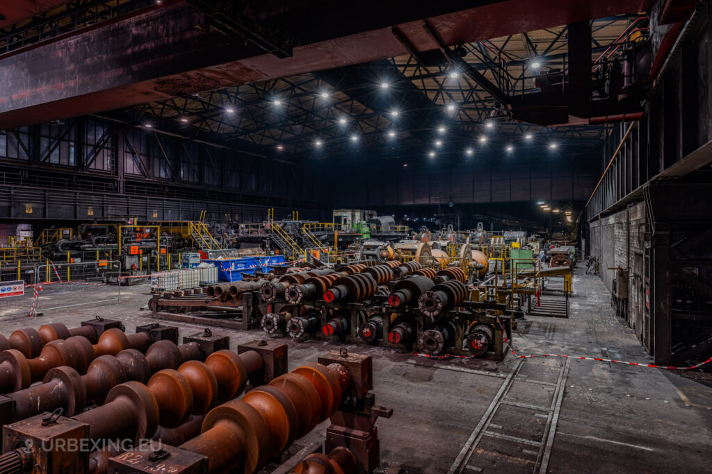 Interior of the Vallourec steel mill in Düsseldorf-Rath, showcasing a variety of rusted industrial rollers, machinery, and metal components under a dimly lit, expansive factory roof.