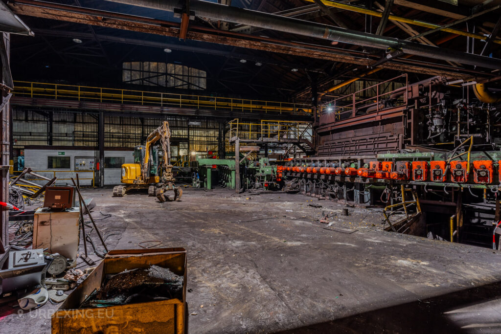 Industrial workshop in the abandoned Vallourec steel mill in Düsseldorf-Rath, featuring a yellow excavator, scattered debris, and rows of orange machinery under a high arched roof.